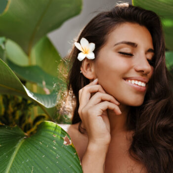 charming woman with white flower dark hair smiles sweetly with closed eyes among tropical tree with large leaves