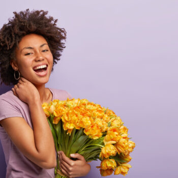 sideways shot glad dark skinned woman laughs with joy touches neck holds yellow tulips wears violet t shirt pleased by getting flowers compliment poses purple wall free space
